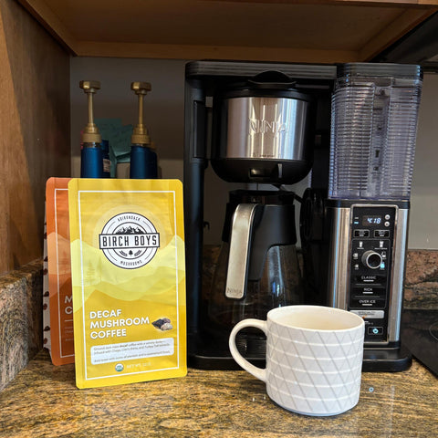 Decaf mushroom coffee package next to a coffee maker and white mug on a kitchen counter.