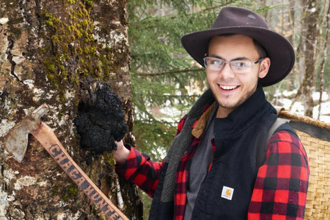 Person in a forest setting with a tree and axe, wearing a hat and plaid jacket.