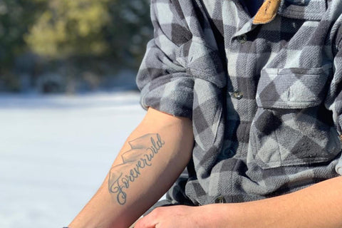 Person wearing a checkered shirt with a tattoo on their arm, sitting outdoors.