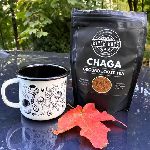 A black stand-up pouch of Birch Boys Chaga Ground Loose Tea stands next to a white enamel mug with black mushroom illustrations, with a red autumn leaf in front, set outdoors on a dark reflective surface with blurred green foliage in the background.