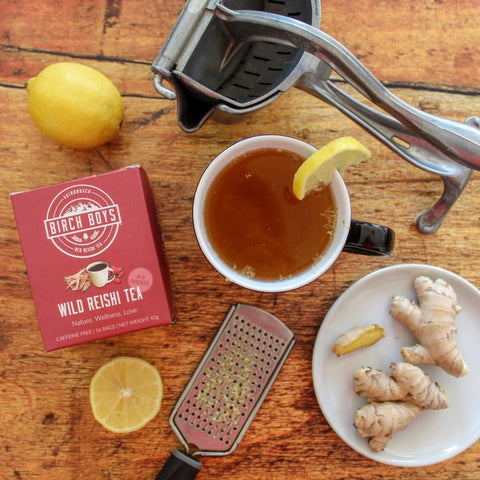 Tea-making setup with a cup of tea, ginger, lemons, and a box of Birch Bots Wild Reishi Tea on a wooden surface.