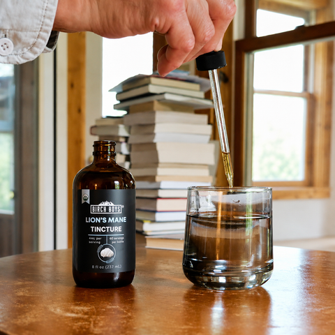 A hand holds a glass dropper above an amber bottle labeled 'Birch Boys Lion's Mane Tincture' while golden liquid is transferred into a clear glass on a wooden table, with a blurred stack of books and window in the background.
